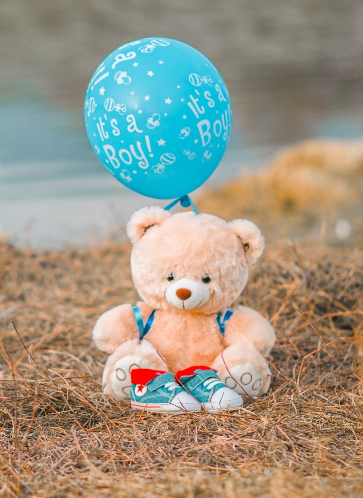 A photo of a teddy bear with a blue "It's a Boy" balloon suspended from its back, and a pair of blue baby shoes in front of it.