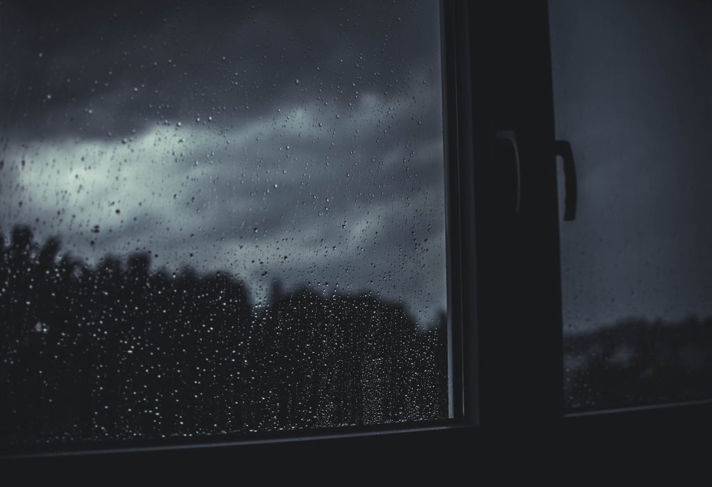A photo of a bedroom window from inside on a stormy night, with rain streaking the window. My age regression story launched on a night like the one in this photo.
