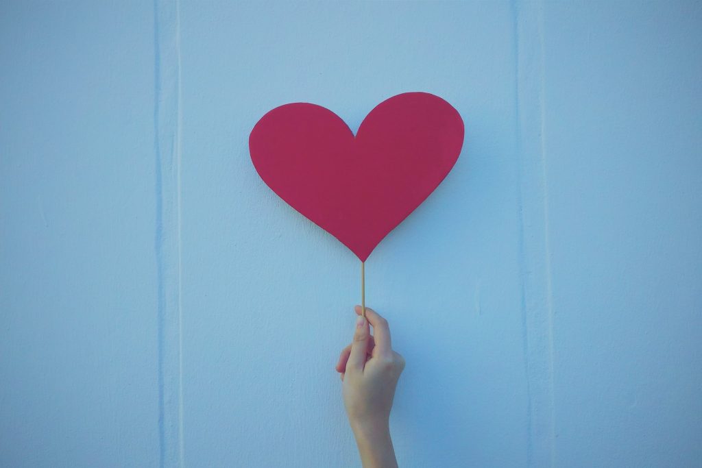 A photo of a hand holding up a pink cutout of a paper heart amidst a blue background.